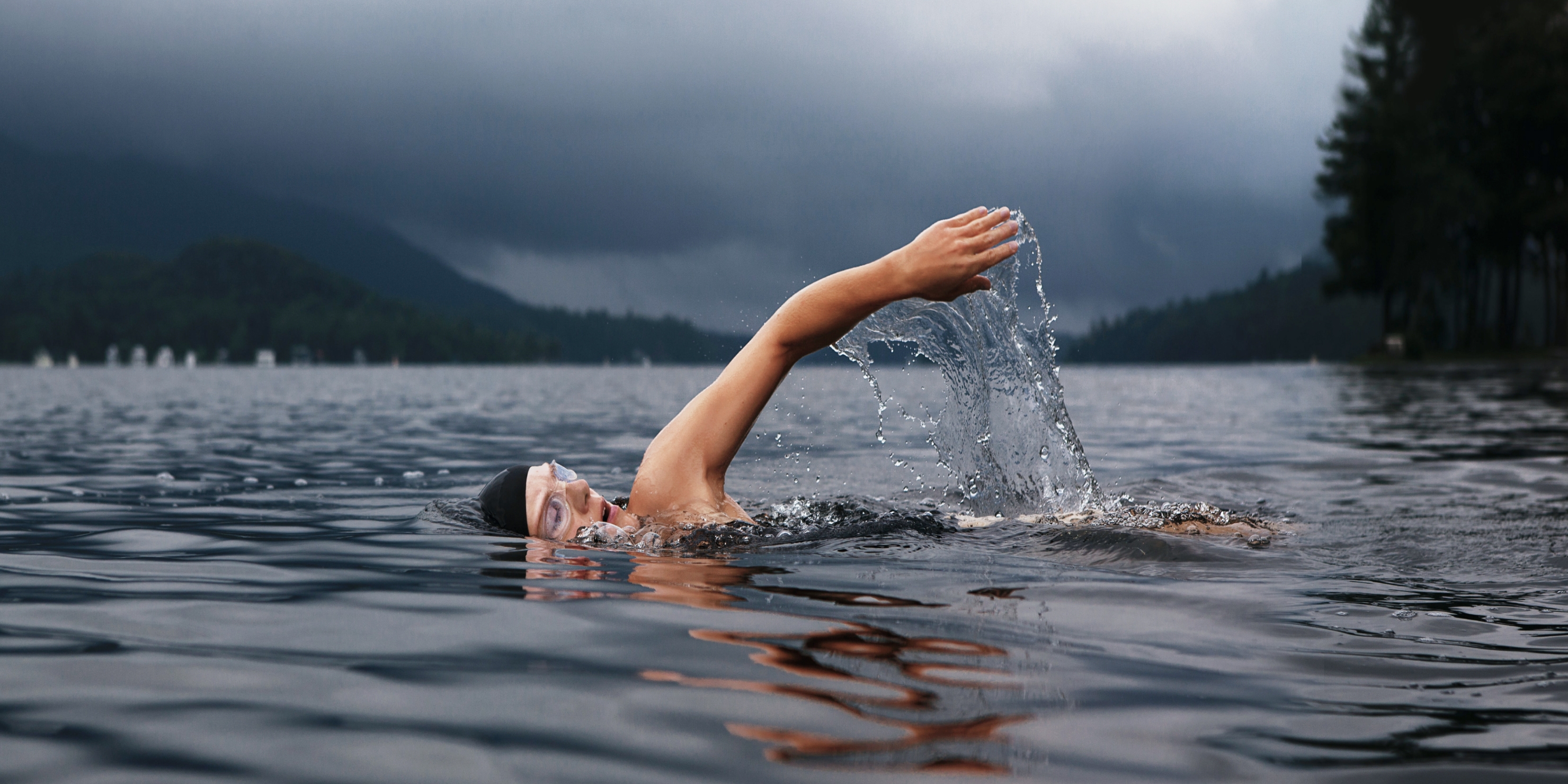 <p>Image of a person front crawling in dark water. Mountains and dark clouds are seen in the background.</p>
